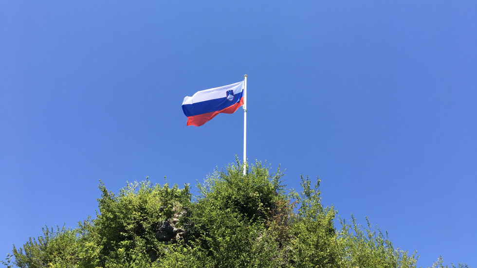 The Slovenian flag on a pole on a hill, fluttering in the wind, with green trees under the flagpole