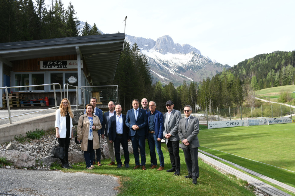 Minister, vodstvo, gneralni konzul, državna sekretarka in drugi udeleženci srečanja stojijo pred igriščem in prostori društva, v ozadju gora. 
