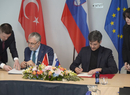 Moška sedita in podpisujeta dokument, na mizi rože, zadaj turška, slovenska in evropska zastava.