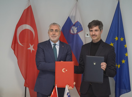 Moška stojita in se rokujeta. V roki držita mapo. Zadaj slovenska, turška in evropska zastava.