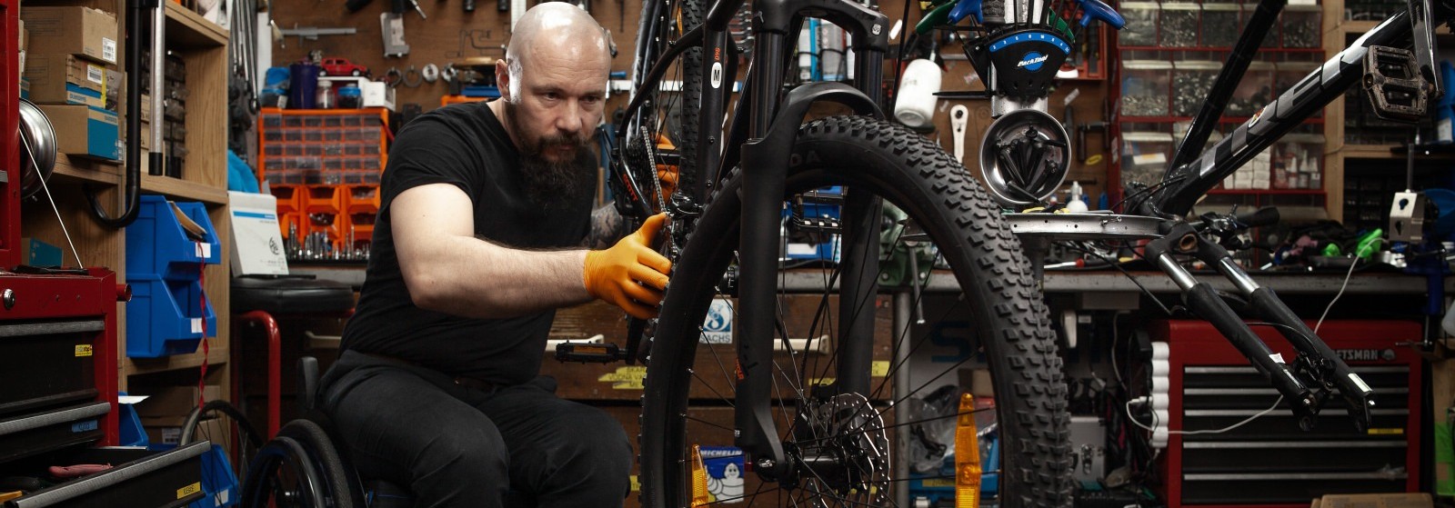 The photo shows a person in a wheelchair repairing a bicycle in a bicycle repair shop.