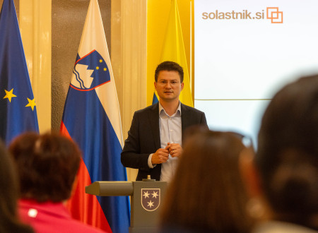 Tilen Božič za govorniškim odrom, zadaj zastave in panoji ter platno