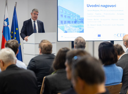 Podpredsednik za govorniškim pultom, zadaj prezentacija, pred njim udeleženci konference