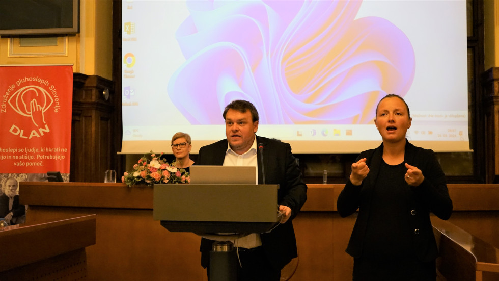 Minister za govorniškim pultom ima nagovor za udeležence posveta, zraven njega stoji tolmačka za znakovni jezik