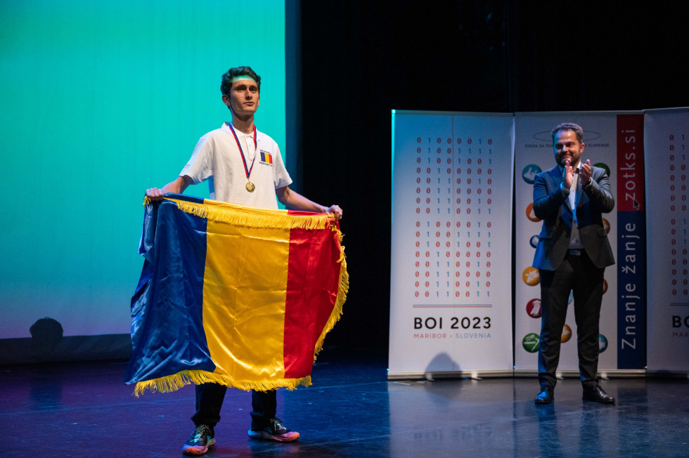 Mladenič z romunsko zastavo in zlato medaljo, za njim pa državni sekretar dr. Miroslav Kranjc, ki mu ploska.