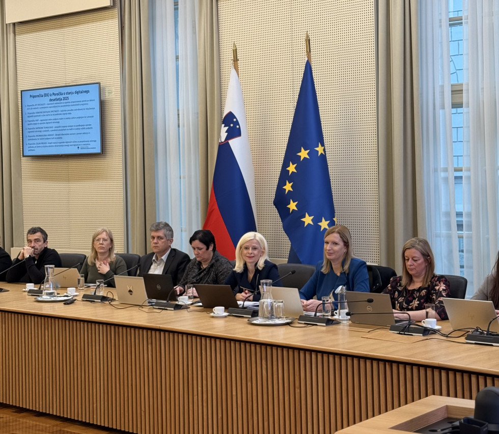 Moški in ženske sedijo. Zadaj slovenska in evropska zastava. 