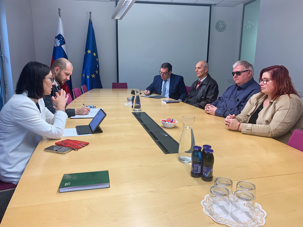 Ministrica dr. Emilija Stojmenova Duh, vodjem Sektorja za digitalno vključenost dr. Tilnom Gorenškom in štirimi predstavniki Zveze slepih in slabovidnih Slovenije sedi med pogovorom za mizo. 