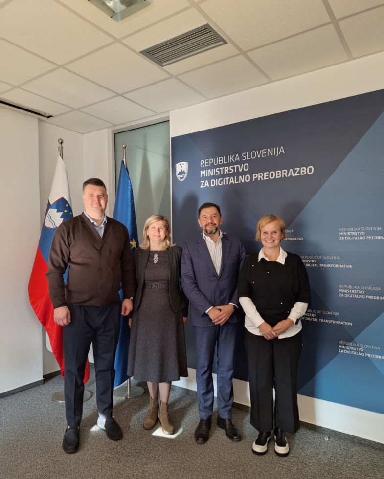 Dva moška in dve ženski stojijo eden ob drugem. Zadaj zastave in velik pano z napisom Ministrstvo za digitalno preobrazbo.