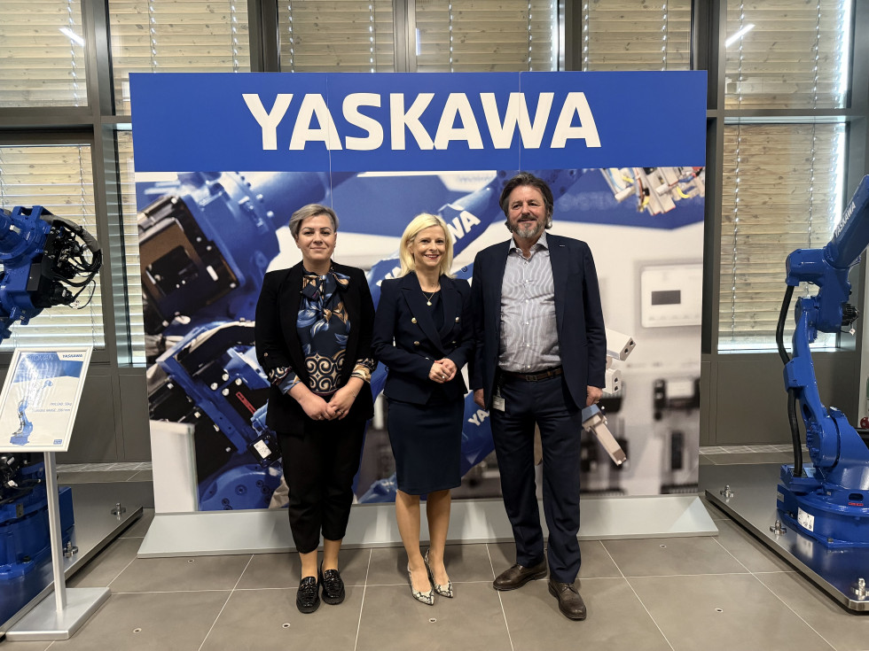 Ministrica, ženska in moški stojijo eden zraven drugega. Zadaj napis: Yaskawa