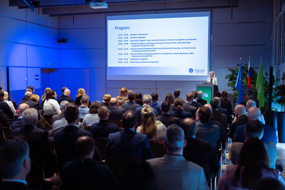 Polna dvorana, ljudje sedijo na stolih. Zadaj na velikem platnu predstavitev. Ženska ima govor za govorniškim pultom.