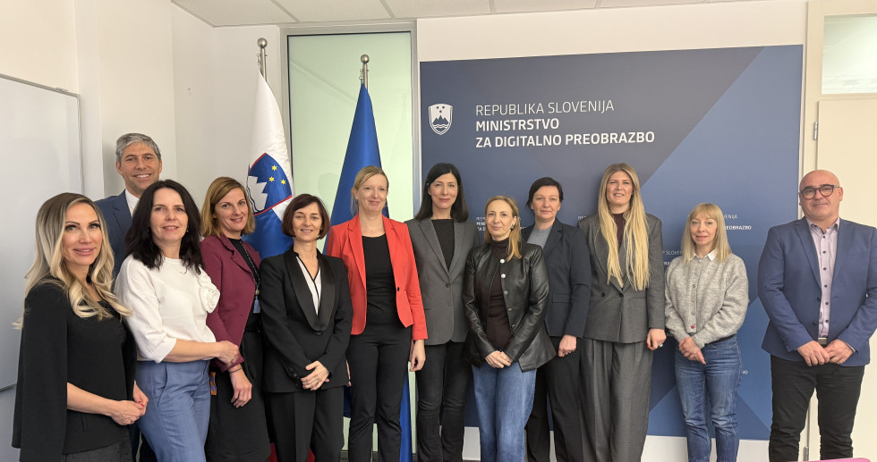 Moška in ženske stojijo eden zraven drugega. Zadaj slovenska in evropska zastava ter pano Ministrstva za digitalno preobrazbo.