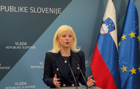 Ministrca Klampfer (A blonde woman in a dark blazer is speaking at a podium during a press conference. Behind her are the flags of Slovenia and the European Union, along with a blue backdrop displaying Slovenian government branding.)