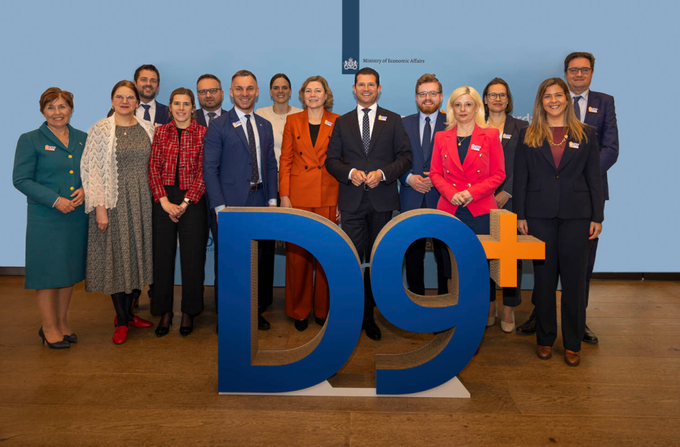 Skupina ljudi v poslovnih oblačilih stoji za velikim znakom "D9+" v konferenčni sobi z modrim ozadjem.