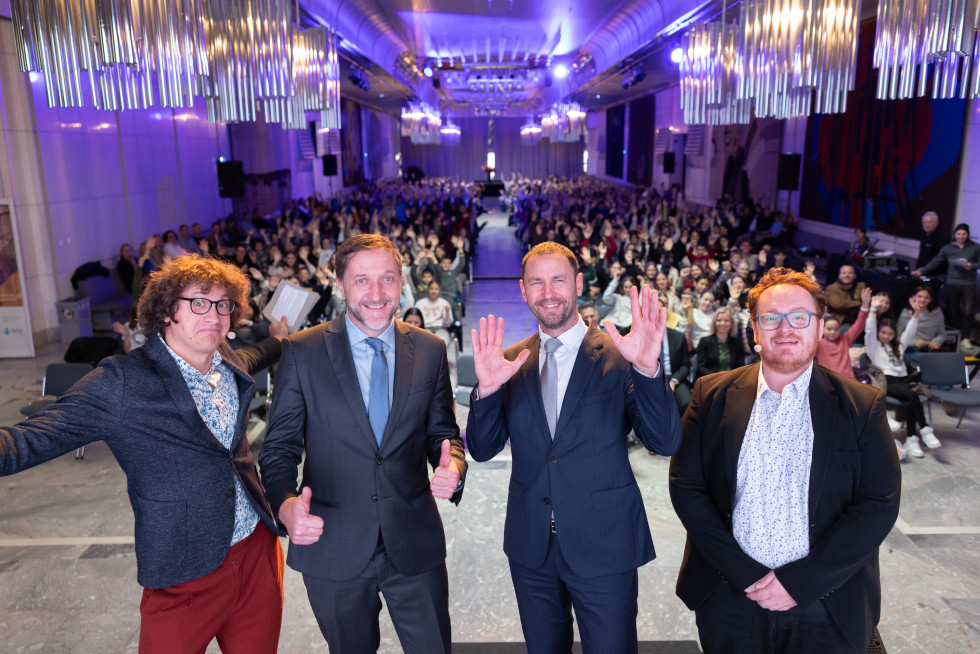 Vsi štirje stojijo na odru in pozirajo, v ozadju sedijo otroci