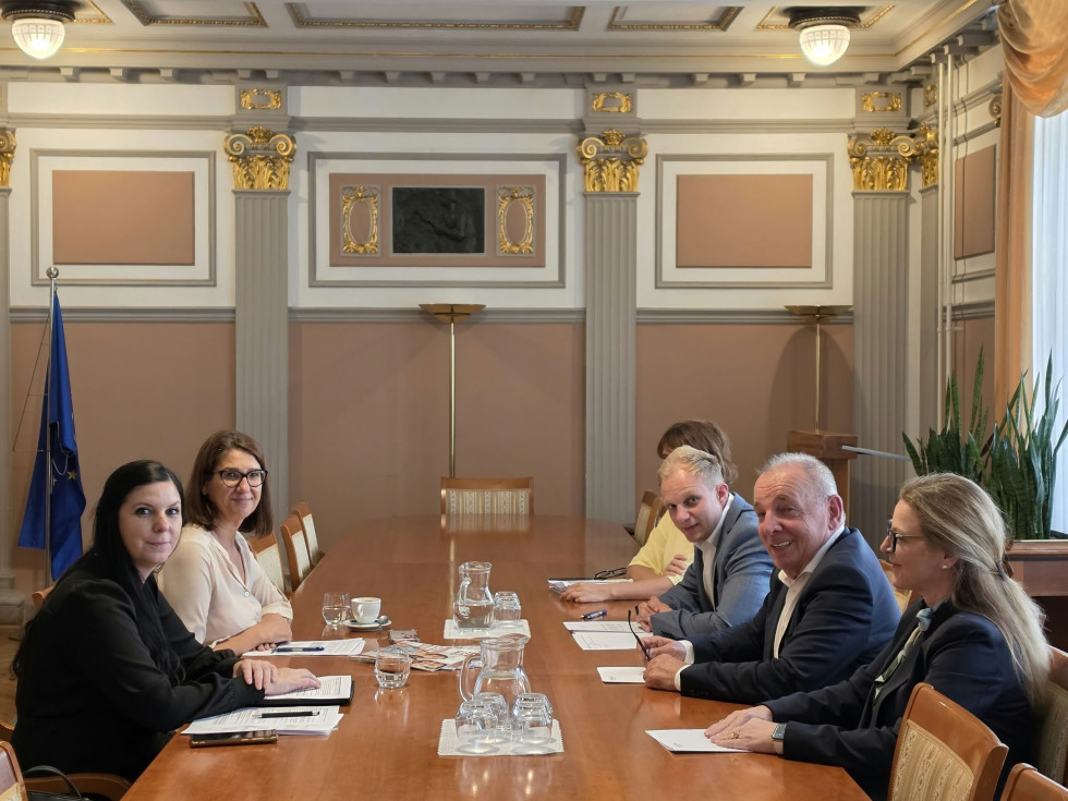 Moški in ženski v poslovnih oblačilih sedijo za mizo, vsi gledajo proti kameri in se smejijo.