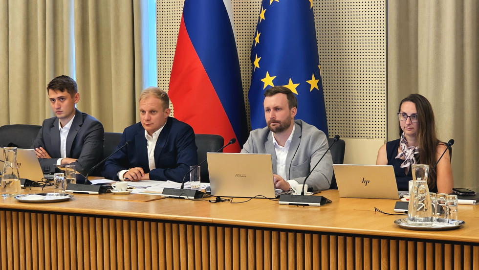 Za mizo sedijo trije moški v oblekah in ena ženska, pred sabo imajo računalnike in zapiske, za njimi sta slivenska in evropska zastava.