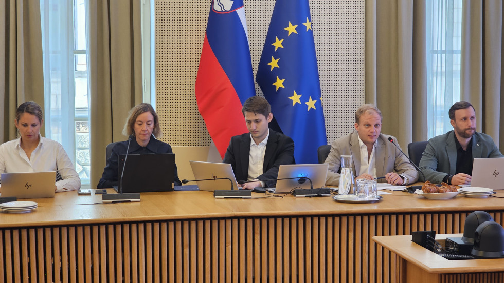 Moški in ženske v poslovnih oblekah sedijo za mizo, pred sabo imajo prenosne računalnike, za njimi pa sta slovenska in evropska zastava.