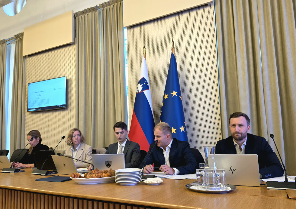Dve ženski in trije moški sedijo za konferenčno mizo z mikrofoni in dokumenti, v ozadju sta slovenska in evropska zastava.