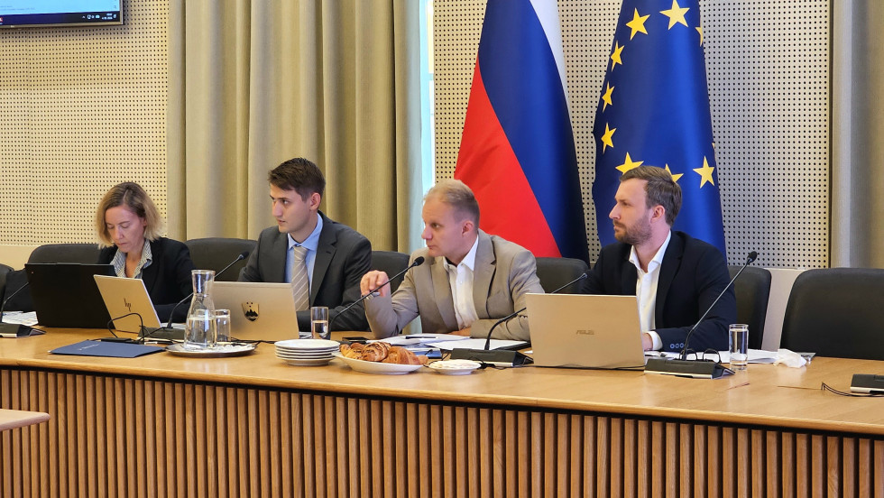 Trije moški in ena ženska v poslovnih oblekah sedijo za dolgo mizo, pred njimi so računalniki in mikrofoni, za njimi pa slovenska in evropska zastava.