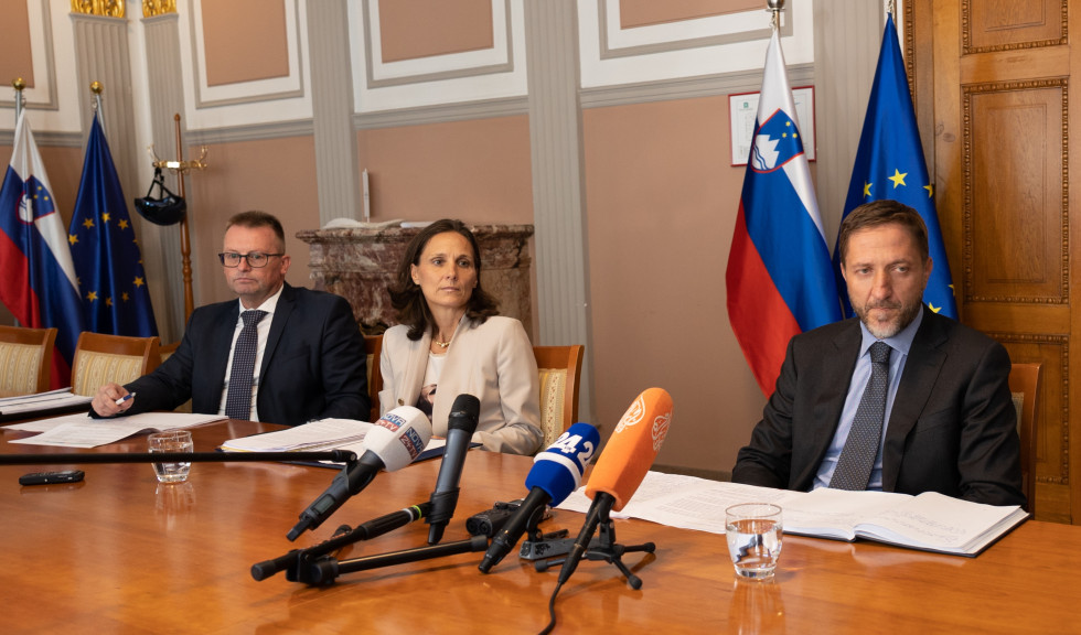 Minister s sodelavcama sedi za mizo in odgovarja na vprašanja novinarjev. Na mizi so mikrofoni, v ozadju pa zastave Slovenije in Evropske unije.