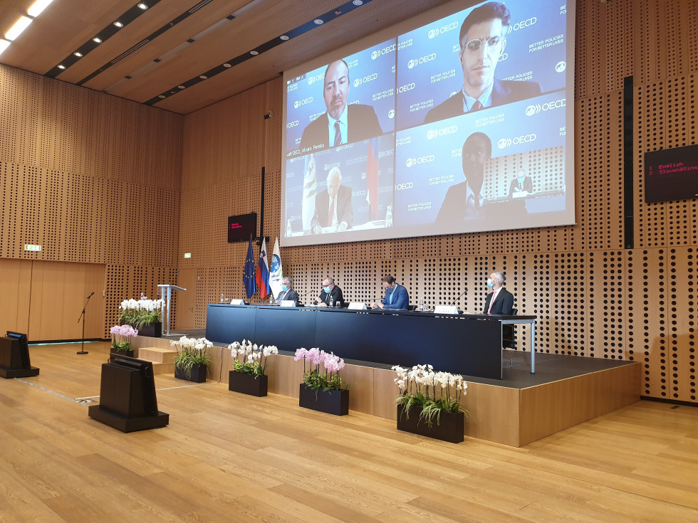 Oder sejne sobe v Kongresnem centru Brdo. Na odru za mizo sedijo (od leve prosti desni) ministri Gantar, Šircelj, Logar in Cigler Kralj. Za njihovim hrbtom na projektnem platnu se preko videokonference oglašajo štirje predstavniki OECD. 