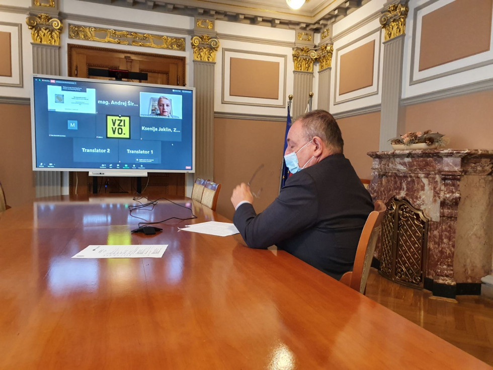 Minister sedi za veliko rjavo mizo v sejni sobi. Levo pred njim velik ekran, na katerem se predvaja videokonferenca. Minister ima pred sabo papirje in na obrazu masko.  