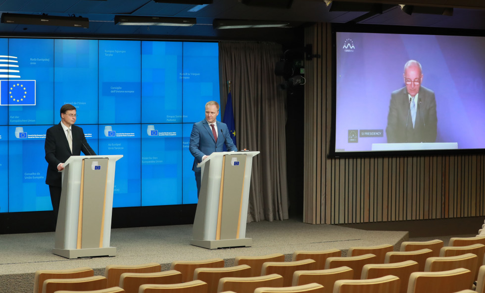 Evropski komisar Valdis Dombrovskis in uradni govorec stojita za govorniškim pultom, zraven njiju pa je velik ekran, preko katerega se na daljavo iz Ljubljane oglaša minister Šircelj