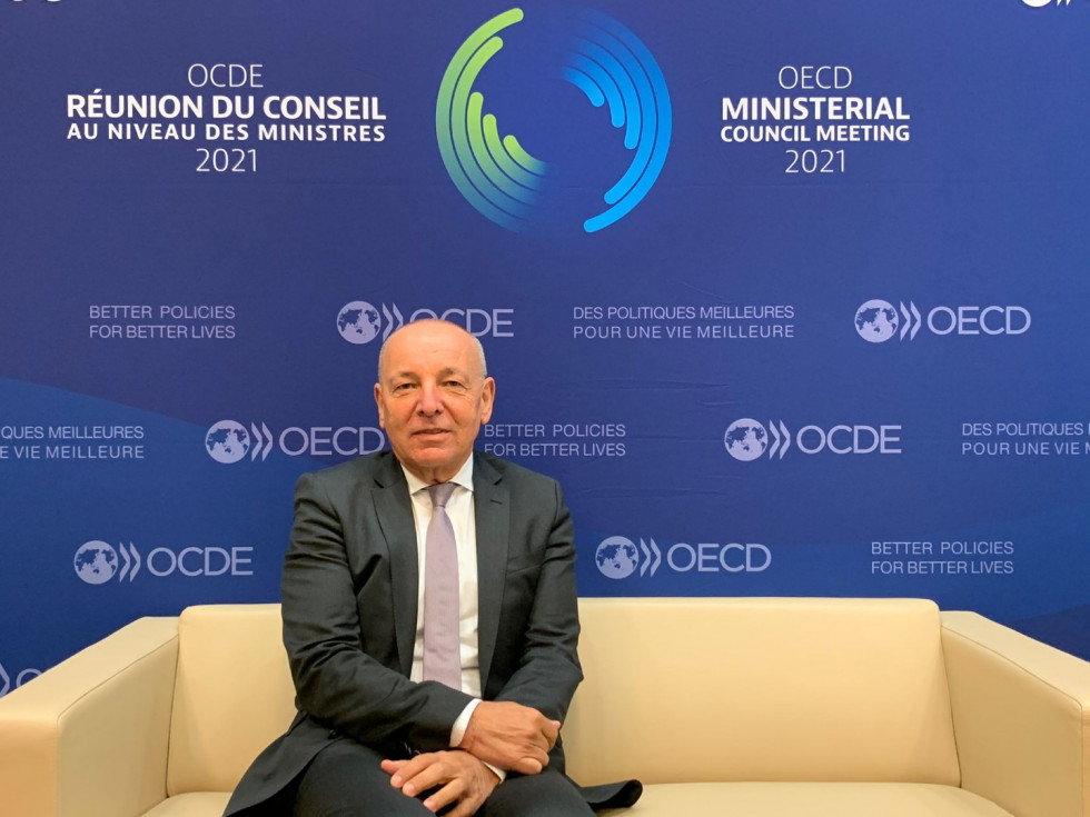 Minister Šircelj sedi na svetlo rumeni zofi. V ozadju na modri podlagi logotip ministrskega srečanja OECD. 