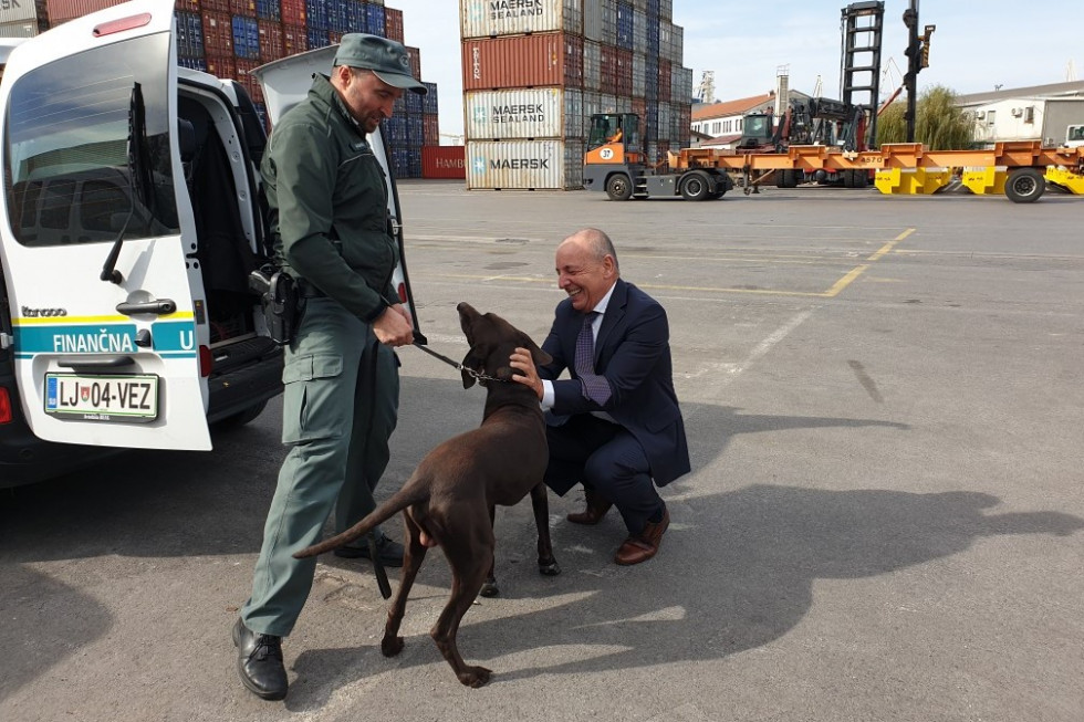 Minister Šircelj v Luki Koper boža službenega psa Finančne uprave po imenu IX. Psa na povodcu drži uslužbenec Finančne uprave v zeleni uniformi. V ozadju ladijski kontejnerji za prevoz tovora. 