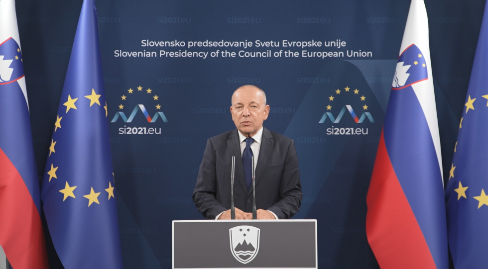 Minister Šircelj stoji pred govornim pultom. V ozadju na modri podlagi logotip predsedovanja Svetu EU. Levo in desno za ministrovim hrbtom evropska in slovenska zastava. a.