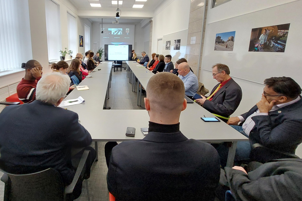 V dvorani sedijo udeleženci dogodka, fotografirani s hrbtne strani, spredaj pred projekcijo sedi minister z moderatorko.
