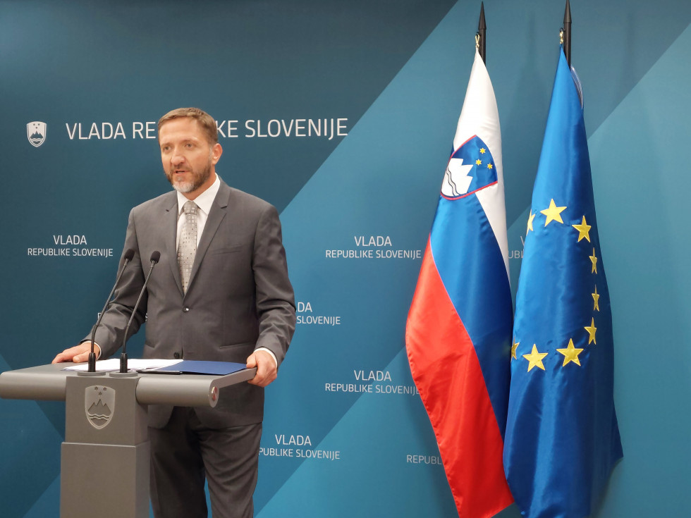 Minister stoji za govorniškim pultom, v ozadju je na steni napis Vlada Republike Slovenije, desno od ministra stojita slovenska in EU zastava 