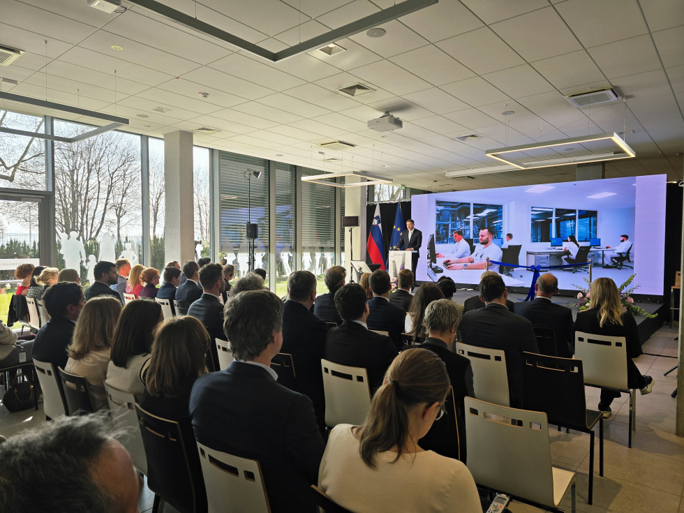 Minster stoji za govorniškim pultom in govori, gosti v dvorani sedijo in poslušajo