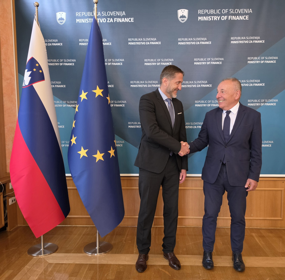 Dve osebi v poslovnih oblekah stojita pred zastavami Slovenije in Evropske unije ter pozirata za fotografijo v uradnem prostoru.