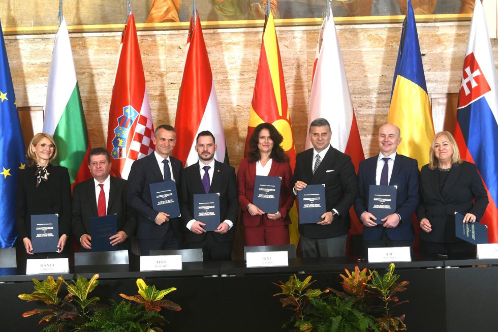 Lepo oblečeni moški in ženske stojijo pred zastavami, v rokah pa držijo podpisane sporazume.