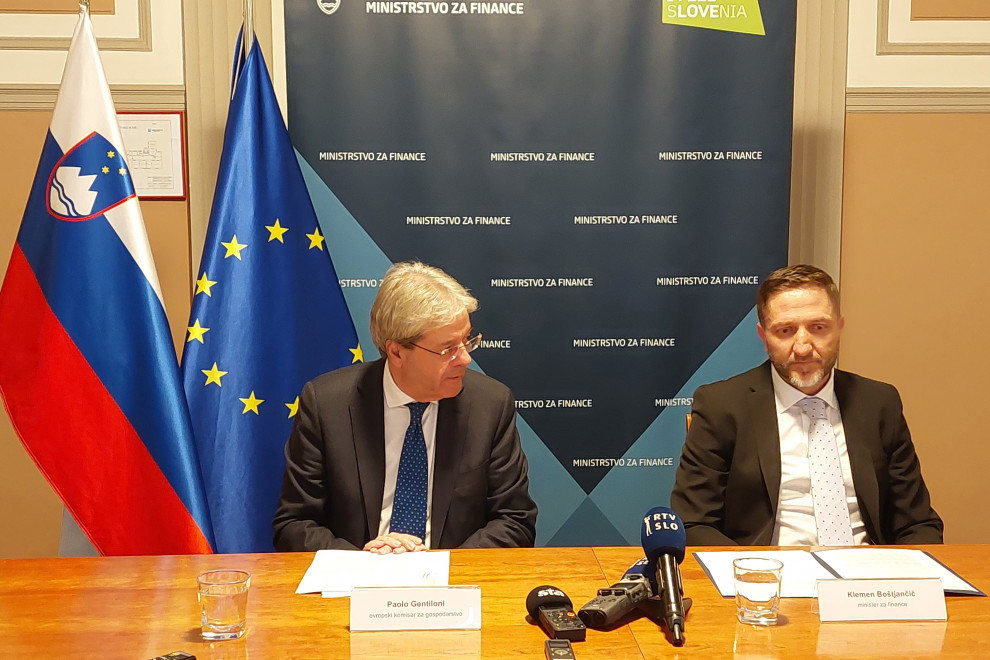 Komisar in minister sedita za mizo, pred njima so mikrofoni. V ozadju sta zastavi Slovenije in EU ter moder pano z napisom Ministrstvo za finance.