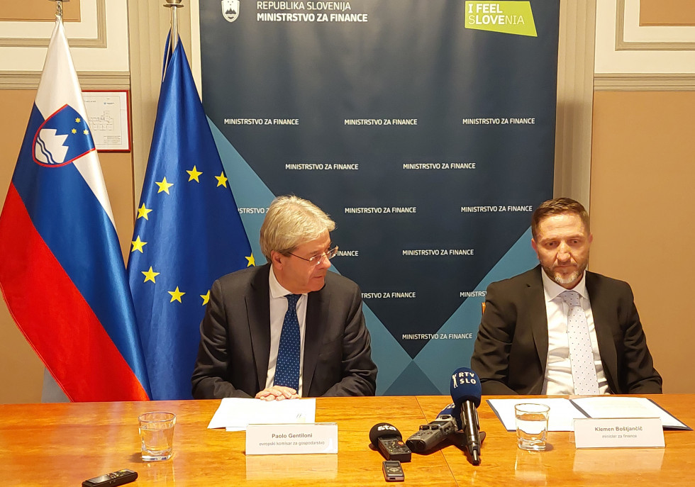 The Commissioner and the Minister are sitting at the table with microphones in front of them. In the background are the flags of Slovenia and the EU and a blue billboard with the sign Ministry of Finance on it.