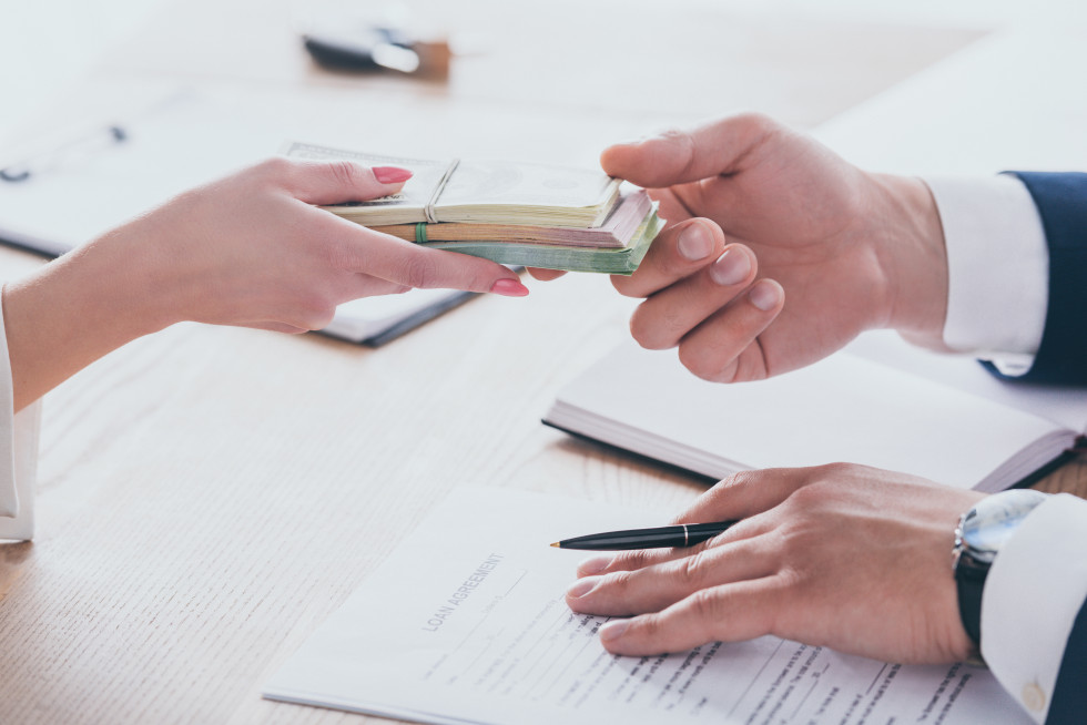 Ženska roka daje moški roki šop denarja, pod rokami na mizi pa so papirji, ki nakazujejo na sklepanje posojila oziroma kredita.