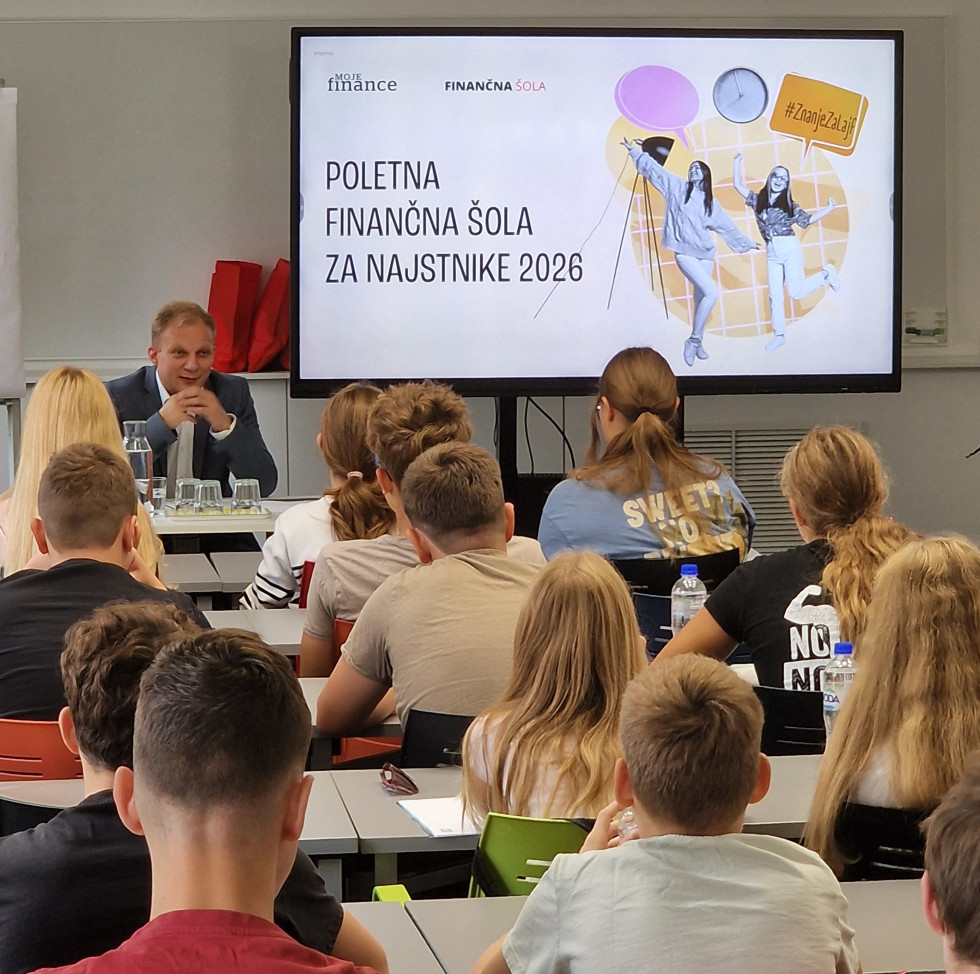 Državni sekretar med nagovorom udeležencev sedi za mizo. Zadaj predstavitev. Učenci sedijo za mizami.