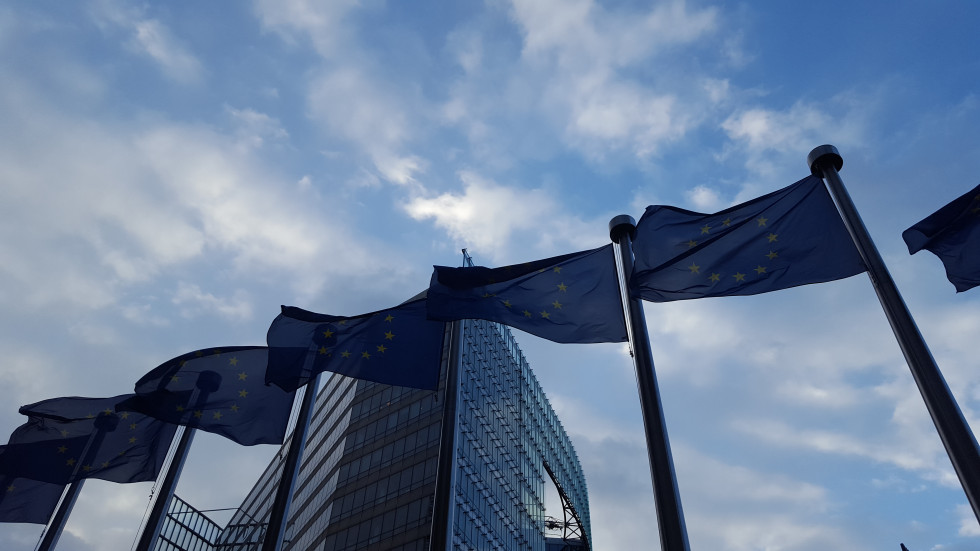 Flags of the European Union fly on poles, fluttering in the wind, with one of the European Institutions' buildings in the background