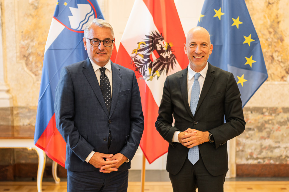 Minister Matjaž Han in avstrijski minister za delo in gospodarstvo dr. Martin Kocher stojita pred slovensko in avstrijsko zastavo. 