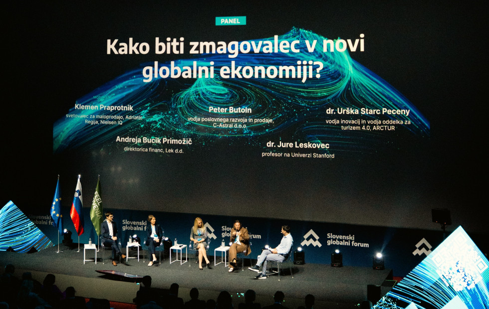 Govorci sedijo na odru in se pogovarjajo. Zadaj velik ekran z izpisanimi njihovimi imeni.