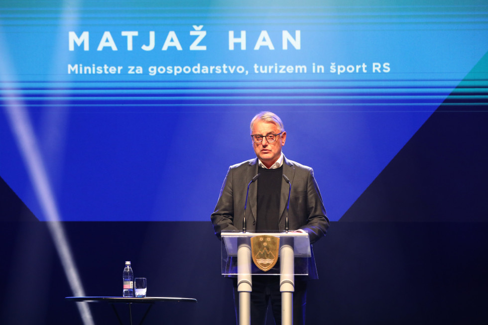 Minister na odru stoji za govornico. Zadaj na velikem ekranu modra podlaga on napisano ime ministra.