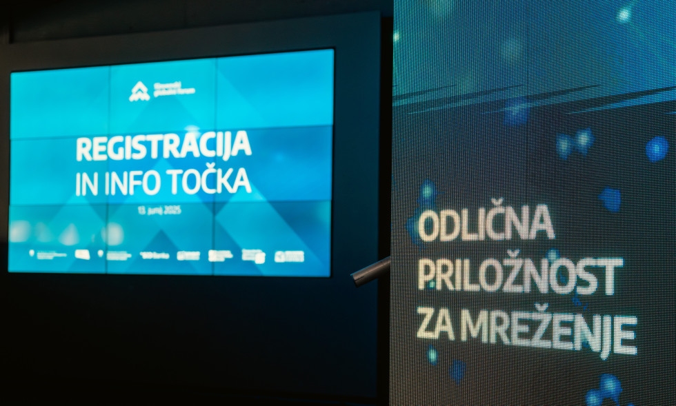 Dva osvetljena zaslona prikazujeta besedilo v zatemnjenem prostoru. Na desnem zaslonu je viden napis: Odlična priložnost za mreženje, na levem: registracija in info točka.