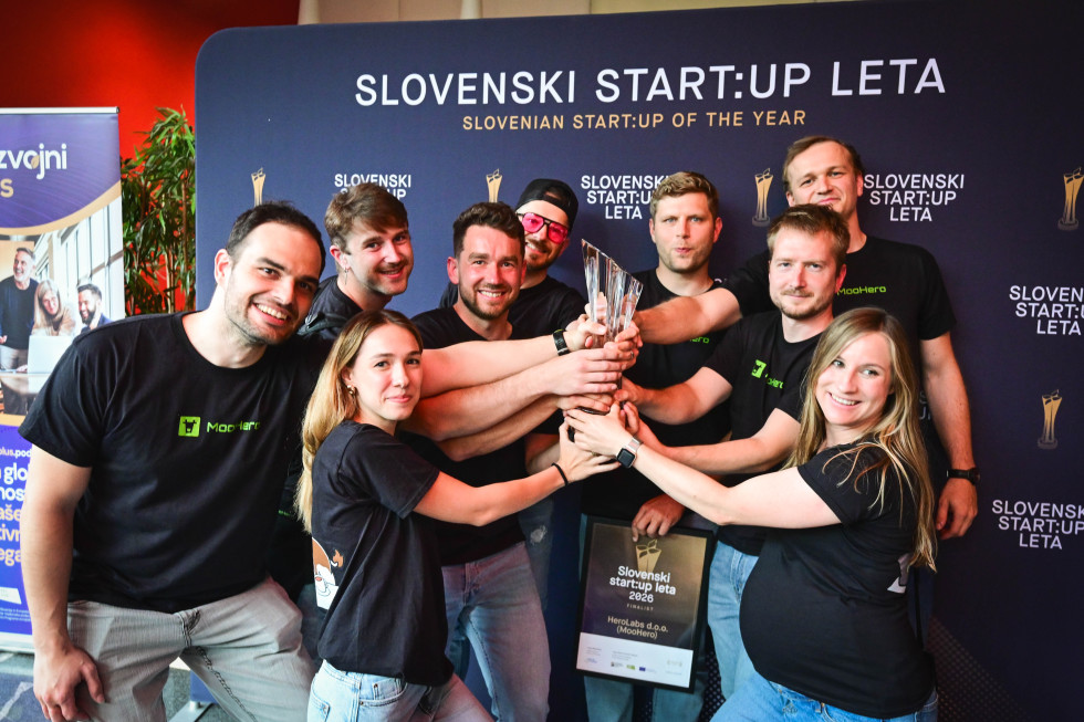 Zmagovalci - skupina oseb stoji na odru in pozira za fotografijo. V sredini skupaj držijo stekleno nagrado, v ozadju pa je viden napis o startupih.