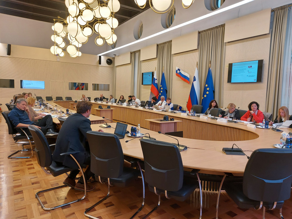 Velika sejna soba z ovalno mizo, zastavami v ozadju in člani sveta ob razpravi