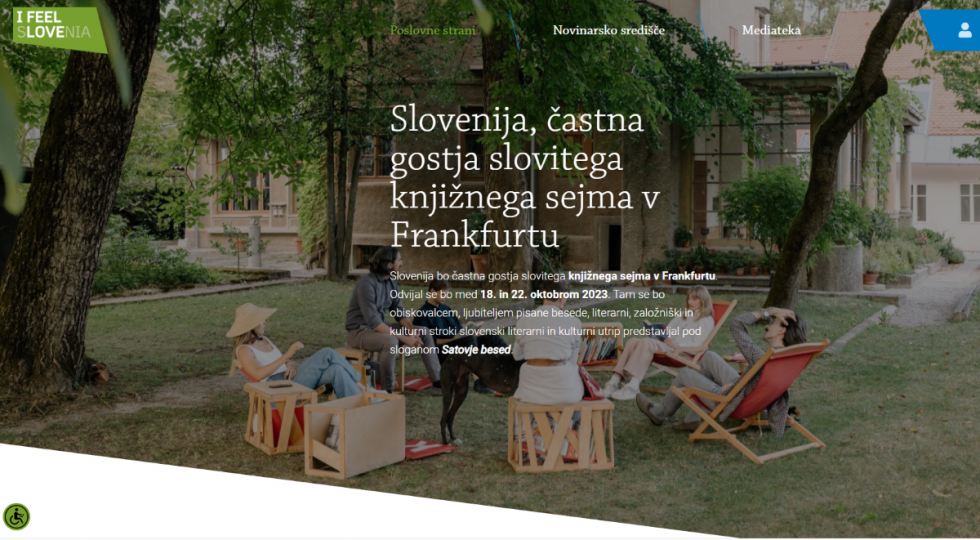 Promocijski banner, ki sporoča, da je Slovenija častna gostja sejma. Na fotografiji je poleg tega zapisa skupina ljudi, ki sedijo na stolih v parku.