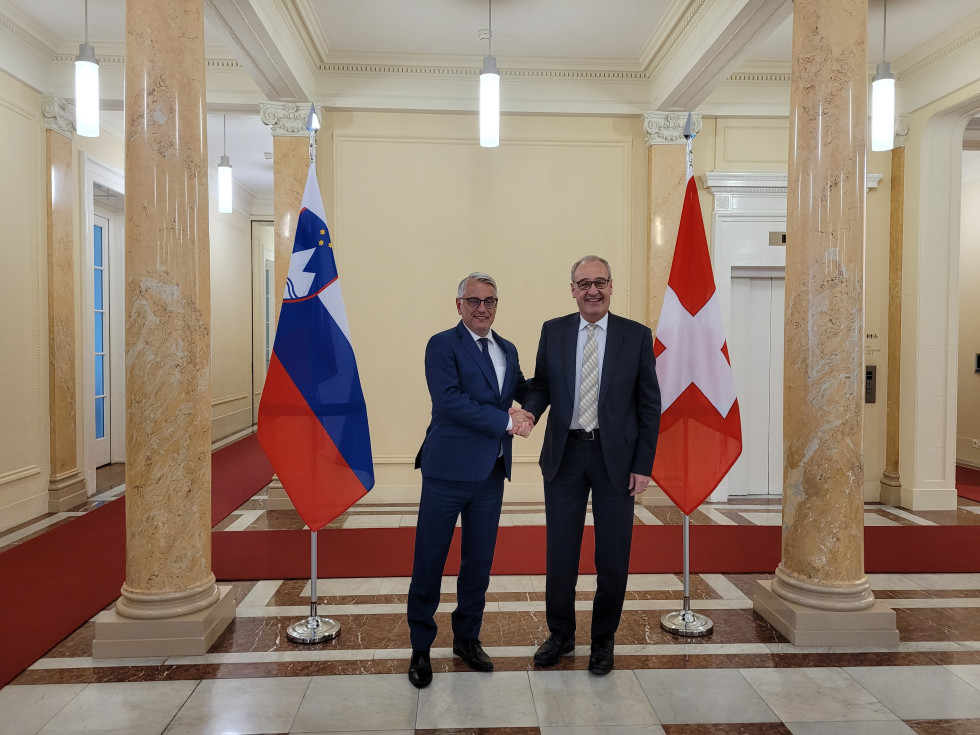 Minister Han in minister Parmelin se rokujeta pred slovensko in švicarsko zastavo. 