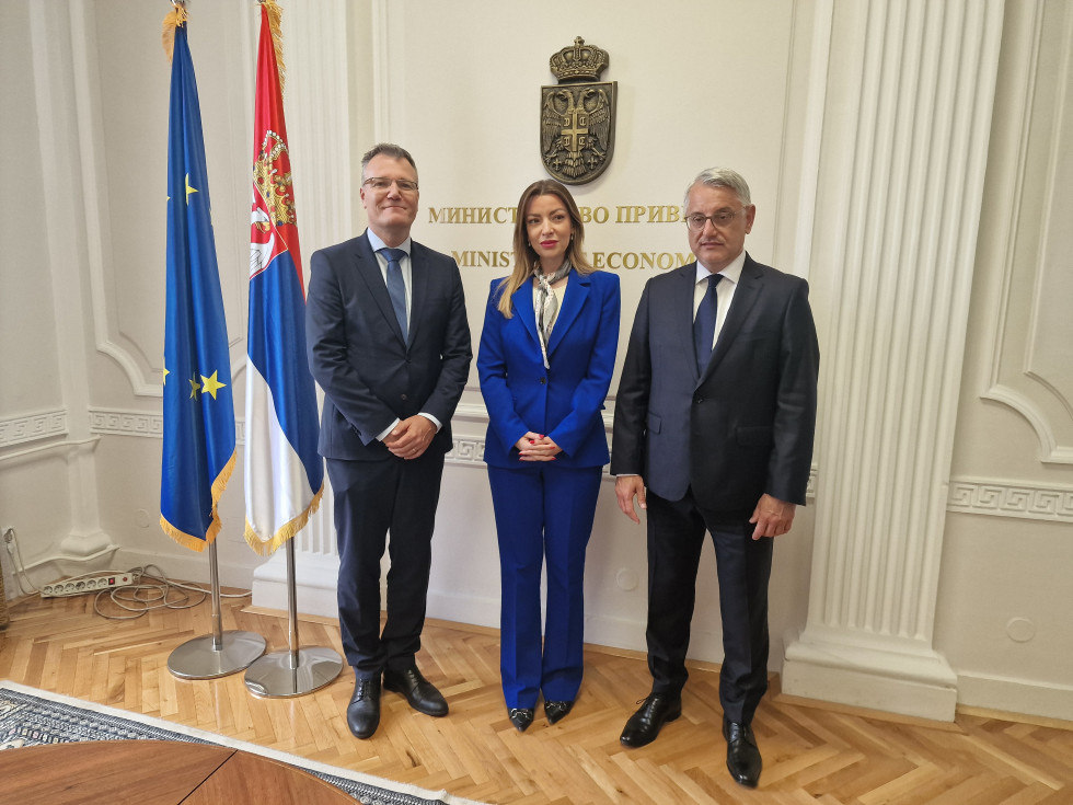 Minister in ministrica skupaj s sodelavcem stojijo ob zastavah, nad njimi na steni srbski grb