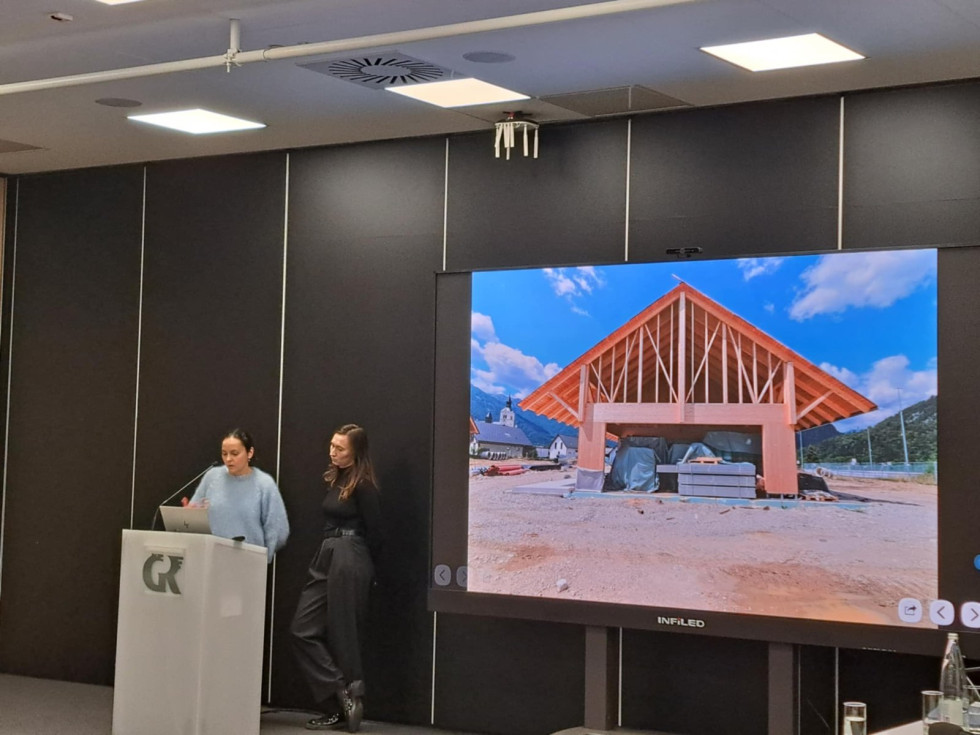 Govornica na odru, v ozadju lesena hiša